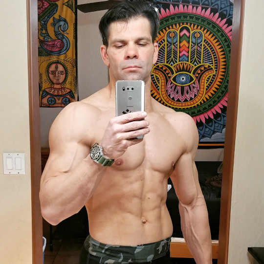 Muscular man taking a shirtless mirror selfie in a decorated room.
